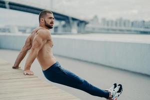 Purposeful bearded adult man trains biceps while doing reverse push up exercises poses with naked muscular torso concentrated away breathes fresh air, likes fitness and sport. Workout outside photo