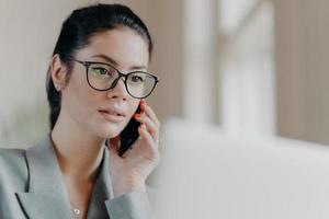 Close up shot of pleasant looking brunette European woman wears transparent glasses, focused in laptop screen, has telephone conversation during remote work, works on project. Technology concept photo