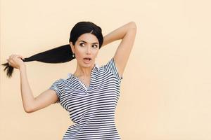 Portrait of attractive brunette female makes pony tail, keeps mouth opened, looks with great interest aside, dressed in striped t shirt, stands on light studio background. Gorgeous lady models indoor photo