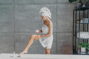 Indoor shot of slim female model applies leg cream stands wrapped in bath towel takes care of body and skin undergoes beauty treatments after taking shower poses in bathroom. Cosmetology concept photo