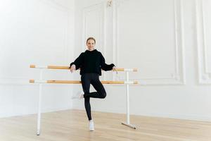 Positive slim woman has dance class, practices in studio near ballet barre, stands on one leg, wears black outfit and white sportshoes, has strong muscles, nice body shape. Gymnastic concept photo