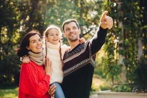 Horizontal portrait of affectionate family spend free time together, enjoy calm atmosphere. Affectionate parents play with their small child, spend weekend in open air. Relationship concept. photo