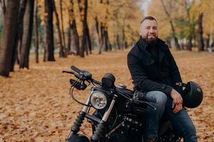 Bearded male motorcyclist rides black bike, holds helmet, has travel on his own transport, poses in park during fall season, looks happily at camera. Carefree biker enjoys journey or trip on vehicle photo