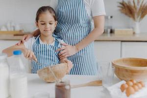 Cropped image of affectionate mother in apron embraces daughter who learns how to make pasty, holds whisk, mixes ingredients in bowl, poses at kitchen table with milk, melted chocolate, eggs photo