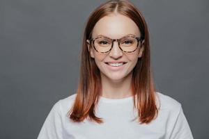 una foto en la cabeza de una joven de pelo oscuro de aspecto agradable lleva gafas transparentes, vestida con ropa informal, tiene una piel sana, un aspecto positivo, muestra dientes blancos perfectos, aislada sobre un fondo gris.