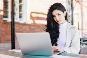 Portrait of beautiful brunette female freelancer typing a message via email using laptop while sitting on terrace looking at camera with confident look. Young lady working with computer outdoors photo