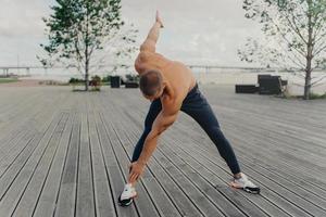 Handsome motivated unshaven sportsman does exercises on promenade, warms up before running, dressed in active wear, poses outdoor. Male runner with naked torso keeps fit, leads healthy lifestyle photo