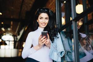 mujer atractiva con cabello oscuro, ojos brillantes y pestañas largas con gafas de sol en la cabeza mirando pensativamente hacia arriba teniendo algunos sueños mientras se comunica con su amante a través de las redes sociales, usando internet foto