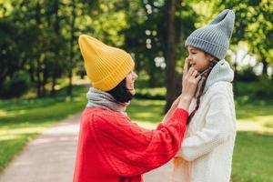 Glad female wears trendy yellow hat and red sweater with scarf touches nose of her small daughter, have walk together in park, play and communicate. People, relationship and togetherness concept photo