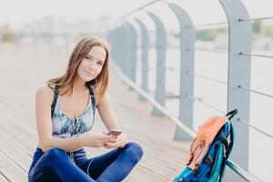 Lovely young woman with fit body, rests after cardio training, dressed in sportswear, holds modern cell phone, listens music in earphones, enjoys perfect sound, her rucksack full of things near photo