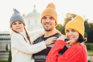 Outdoor portrait of friendly family stand close to each other, have broad smiles. Pretty woman in yellow trendy hat and red sweater, handsome young man holds little adorable daughter on hands photo