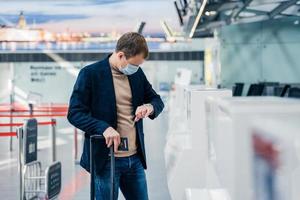 Horizontal shot of man passenger wears disposable medical mask to prevent virus, checks time on his watch, has hour for departure, poses in internation airport with suitcase and passport. Coronavirus photo