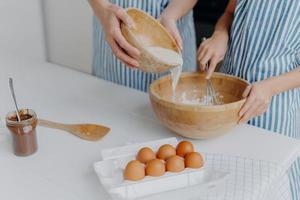 Unrecognizable mother and daughter add milk in bowl, make dough, homemade cookies wear aprons, stand on table with eggs, chocolate and wooden spatula, try family recipe, demonstrate cooking abilities photo