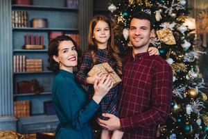 la familia feliz se une contra el abeto decorado, mira felizmente a la cámara, sostiene el regalo, celebra el año nuevo en el círculo familiar. niña pequeña que se alegra de pasar las vacaciones con su madre y su padre foto