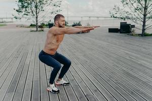 Full length shot of active man does squatting exercise poses with naked torso has muscular body poses outside. Strong athletic fit man in sportswear exercises in open air. Confident fitness instructor photo