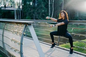 Motivated brunette active young woman does squat exercises with dumbbells, trains biceps, dressed in black active wear, poses at bridge outdoor during sunrise, has morning workout. Sport concept photo