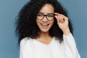 Close up shot of cheerful woman wears glasses, keeps hand on frame, looks gladfully aside, notices something awesome in distance, dressed in casual white jumper, models against blue background photo