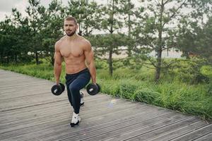 el hombre barbudo motivado con el torso desnudo se pone en cuclillas con pesas pesadas, hace ejercicios físicos al aire libre, disfruta del entrenamiento, posa cerca de árboles verdes, levantando pesas. concepto de estilo de vida saludable foto