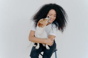 linda mujer feliz con el pelo rizado recibe un beso de jack russell terrier siente amor por su mascota favorita se complace en compañía de un perro sentado en una silla contra un fondo blanco. amor entre dueño y animal. foto