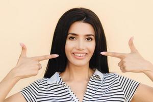 People, beauty and advertising concept. Brunette young woman with gentle smile, indicates at mouth with broad smile, has braces, wears casual striped t shirt, isolated over light studio background photo