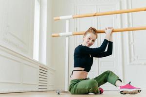 Shot of beautiful ginger woman does pilates exercises on handrail, sits on floor, stretches herself, poses in dancing studio, being in good body shape, dressed in sportswear. Sport and health care photo