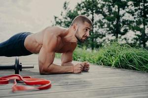 Fitness bearded Caucasian man with naked torso stands in plank pose, exercises outdoor, poses near sport equipment, has strong arms, demonstrates endurance and motivation. Abdominal workout. photo