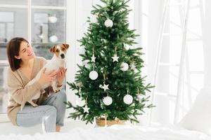 Smiling woman embraces pet with love, has happy mood, pose together near beautiful decorated Christmas tree, white walls, ladder and window around, spend time at home. Winter time, holidays. photo