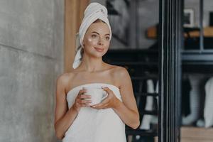 Thoughtful lovely young female model with cream on face, wrapped in towel, concentrated somewhere aside, holds mug of tea, undergoes beauty treatments at home, stands indoor. Body care concept photo