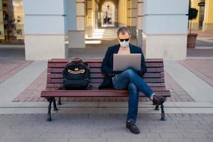 Photo of busy man freelancer in mask does distance work, keyborads on laptop computer, sits at wooden bench outside, avoids crowded places during quarantine time. Traveling and coronavirus concept