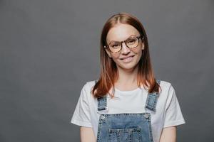 Isolated shot of positive pretty woman tilts head, has charming smile, wears glasses, white t shirt and overalls, poses over grey background with free space for your advertising content or promotion photo