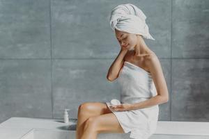 Refreshed young European female applies anti wrinkle cream, poses in bathroom, wrapped in bath towels, prevents signs of skin aging, has clean body after showering. Wellness, wellbeing concept photo