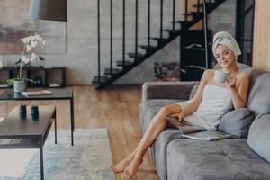 Happy young woman poses wrapped in bath towel, drinks aromatic coffee and reads magazine, sits on comfortable sofa, poses in living room at home. Domestic atmoshpere, leisure time, lifestyle photo