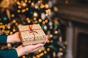 Hirizontal shot of beautiful wrapped present box in womans hands against decorated Christmas or New Year tree. New Year background. Focus on gift box. Celebration concept. photo