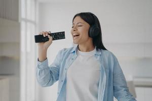 Excited woman in headphones sing to imaginary microphone, TV remote control, listen to music at home photo