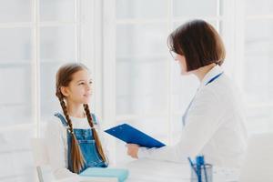 Female pediatrician examines little child, listens carefully to small kid patient, writes down notes in clipboard, pose in clinic or hospital against white window. Children healthcare concept