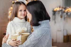 Young female embraces her little child, gives present, congratulates with New Year, proud to have such beautiful and clever daughter. Happy kid spends time with mother. Childhood and holidays photo