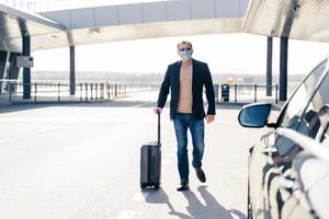 Business traveler pulls suitcase from modern airport terminal, walks to car, wears medical disposable mask on face, prevents from coronavirus during dangerous pandemic situation. Full length shot photo