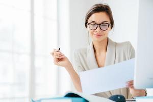 Concentrated successful businesswoman looks attentively in paper, studies terms of contract, holds pen, writes in documentations, dressed formally, poses at desktop against white spacious interior photo