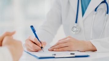 Unrecognizable woman doctor writes down in clipboard, makes prescription for patient, records data for analysis, wears white gown. Close up, focus on hands. Medicine, insurance, health care concept photo