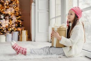 Sideways portrait of restful female child wears white sweater, trousers and warm socks, embraces wrapped gift, sits on floor in cozy room, admires New Year tree. Children and holidays concept photo