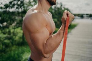 Cropped shot of unrecognizable muscular man has workout biceps exercise stands in profile shows strong muscles, uses resistance band, strives for strong body. Bodybuilding, motivation concept photo