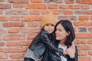 Affectionate mother with dark hair recieves warm hug from daughter, wear leather jackets, pose together against brick wall. Fashionable little girl in hat embraces her mum. Family and children concept photo