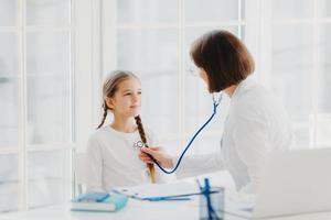 Woman pediatrician gives consultation to small girl, talk about health and symptoms, listens heart with phonendoscope, makes prescription, pose in hospital office. Childrean medical insurance and care photo
