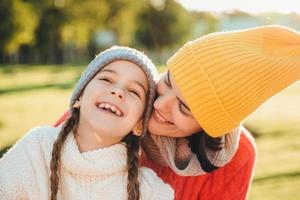 Beautiful female in yellow knitted hat kisses her adorable little child, have wonderful unforgettable time together. Small child smiles happily while recieves kiss from mother. Family relationships photo