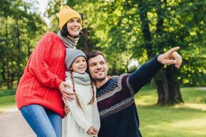 Three family members spend time together, look at beautiful lake in park, indicate with fingers, being in good mood, smile pleasantly. Father, mother and daughter enjoy togetherness, calm atmosphere photo