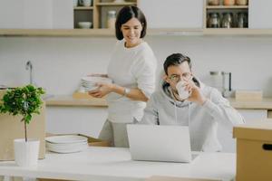 Busy husband works on laptop computer and drinks coffee, wears spectacles and casual sweatshirt, wife busy unpacking belongings, carries dishes, pose in kitchen during moving day. Family and mortgage photo