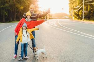 retrato al aire libre de una mujer bonita, su esposo y su hija muestran un buen cartel, caminan con un perro en la carretera, disfrutan del sol, tienen un estilo de vida activo. agradable paseo familiar antes de dormir respirar aire fresco en el campo foto