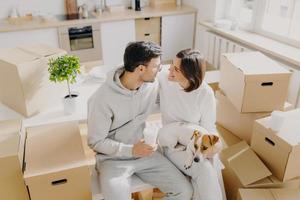 Smiling couple express romantic good feelings to each other, just arrived in new house, pose around cardboard containers, drink takeaway coffee, kitchen furnture in background, dog on womans hands photo