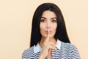 concepto de personas y silencio. mujer atractiva con signo de silencio mantiene el dedo índice en los labios, pide silencio mientras alguien duerme, vestida con ropa informal, gestos en el interior. gente y conspiracion foto