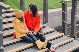 Image of fashionable young woman wears red looe sweater, looks positively at small child, pose together at steps outdoor, enjoy spare time, stroll in urban setting. People, relationship concept photo